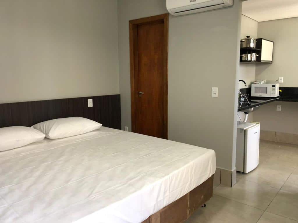 Quarto no Vila Nóbrega Residencial. Uma cama box de casal está na esquerda, e na direita vemos a cozinha de relance. É possível identificar uma pia, micro-ondas, frigobar e armário. Está ilustrando o post sobre airbnb em Foz do Iguaçu.
