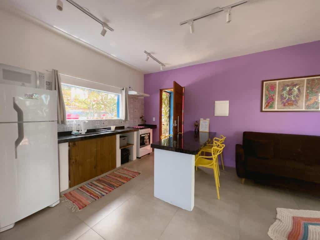 Cozinha e sala do Vila Real Apartamentos. A cozinha está à esquerda, equipada com geladeira, pia, armários, fogão e forno. Há uma bancada com cadeiras no meio. Na direita está o sofá da sala. O ambiente é espaçoso.