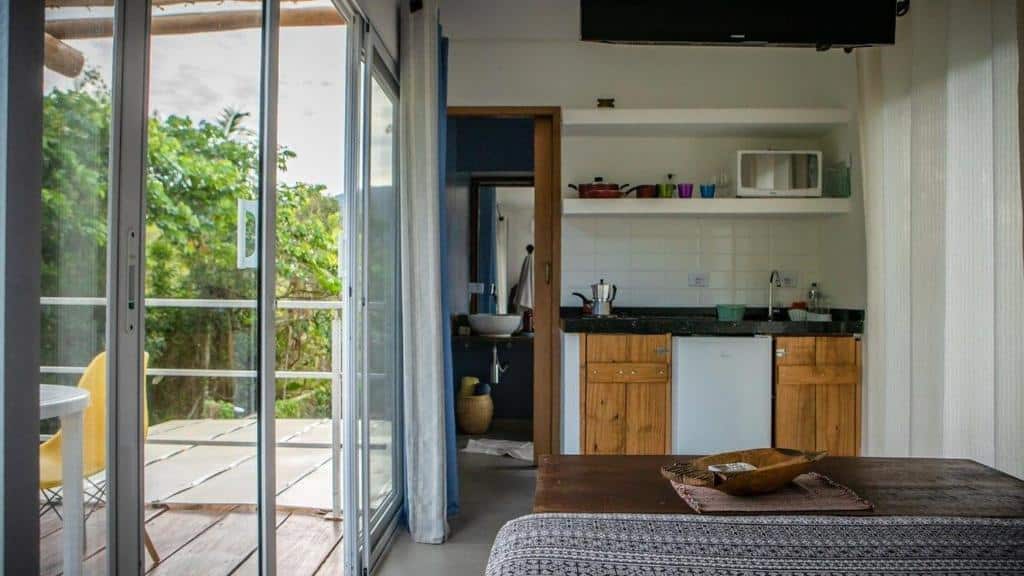 Loft Superior Villa Dallê. Uma cama de casal do lado direito, de frente um baú e uma pia da cozinha com frigobar e micro-ondas, no meio a porta do banheiro, do lado esquerdo a porta de vidro da varanda com uma mesa e cadeiras fora.