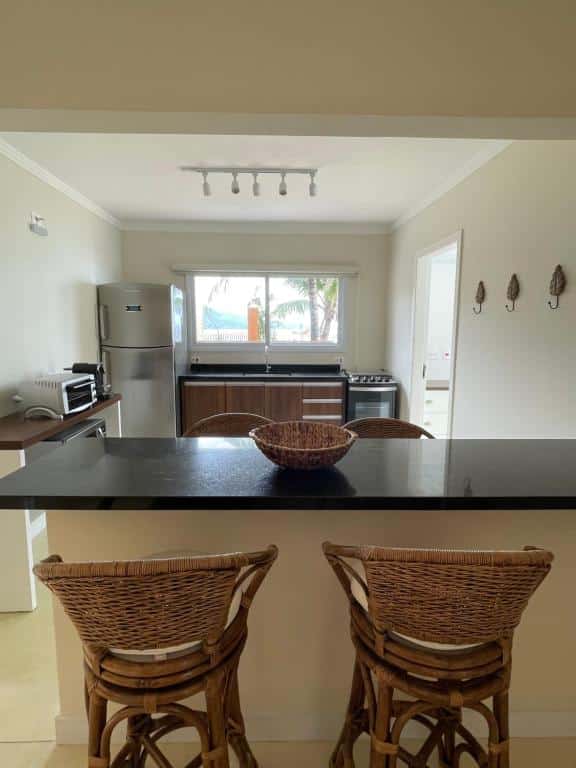 Visão da bancada para a cozinha da Villa di Fiori. Ao fundo estão uma geladeira ladeada pela bancada da pia, e ao lado direto da pia está o fogão. Já ao lado esquerdo da direito fica uma bancada com micro-ondas e cafeteira.