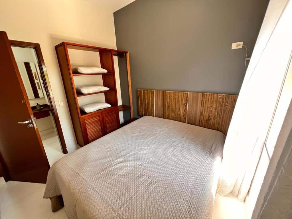 Quarto do Vilaggio Valentina, um dos airbnb em Barequeçaba. Uma cama coberta por lençol está encostada na parede e tem um armário com travesseiros ao lado esquerdo. Uma porta aberta mostra o banheiro logo ao lado do armário. Há uma janela com cortinas do lado direito da cama.