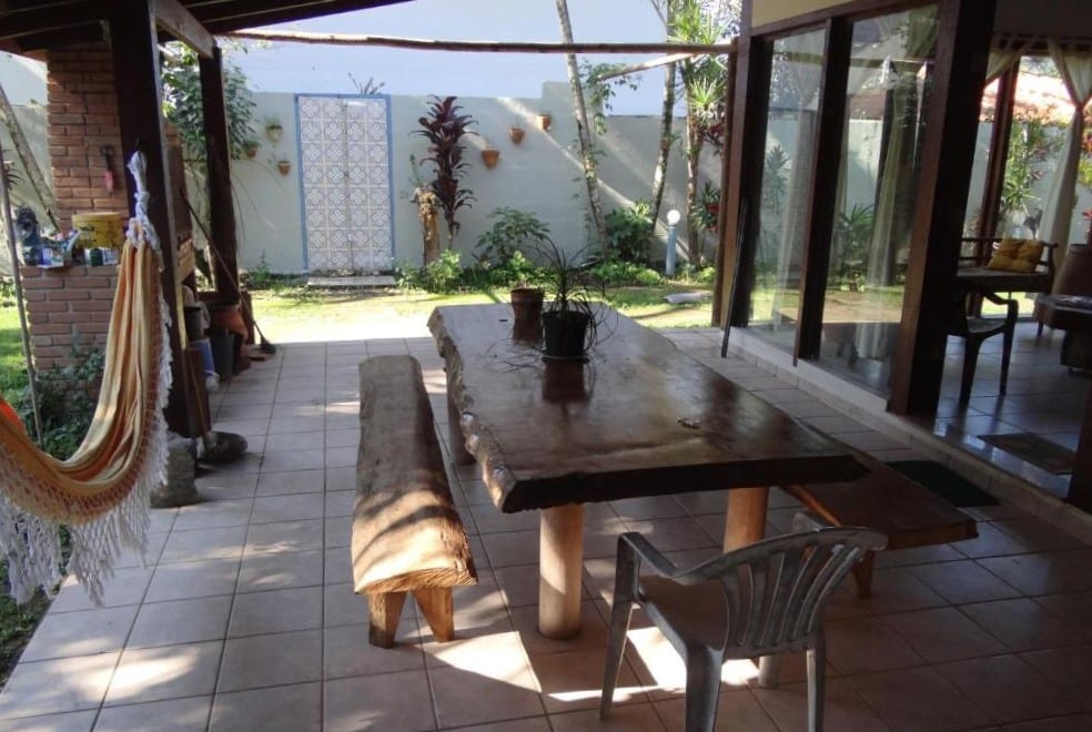 Área externa do Viver Ubatuba. Uma mesa de madeira com um banco de cada lado no meio, do lado esquerdo uma rede e uma churrasqueira, do lado direito a casa.