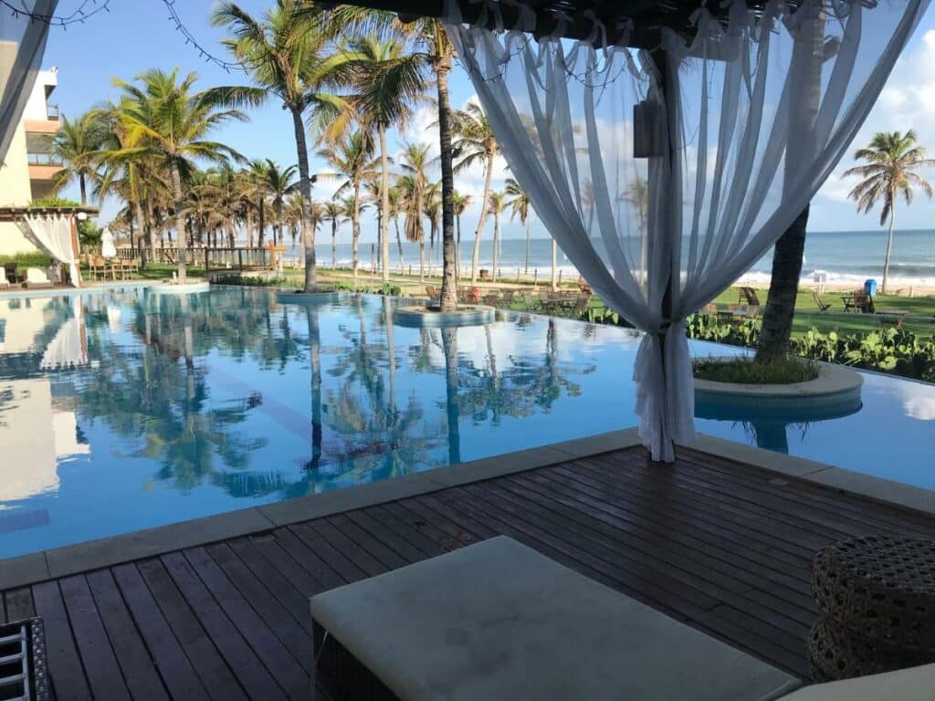 Piscina do resort do airbnb Wai Wai Cumbuco Residence. Ela tem borda infinita, está de frente para a praia e cercado por várias árvores. Um deck de madeira está na borda da piscina. 