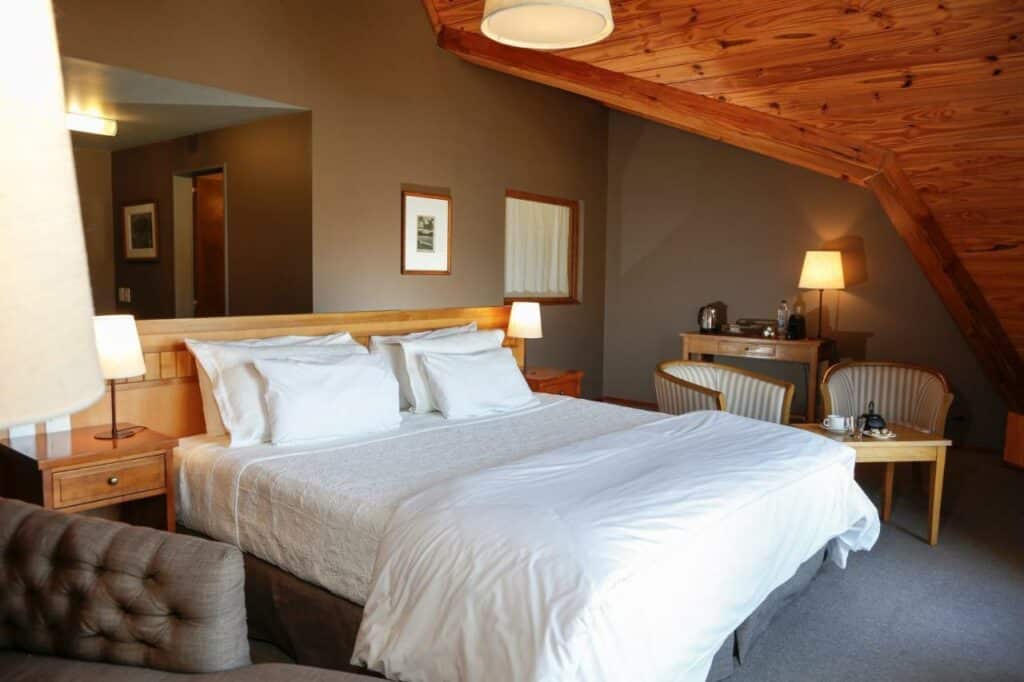 Quarto do Los Cerros del Chaltén Boutique Hotel com cama de casal do lado esquerdo da imagem com uma cômoda em cada lado com luminária e do lado esquerdo da cama duas cadeiras com mesa, para representar hotéis em El Chalten.