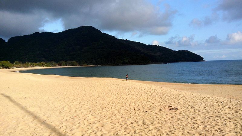 Praia em São Sebastião. A areia é clara e há uma pessoa caminhando por ela,. O mar e algumas montanhas estão no fundo.