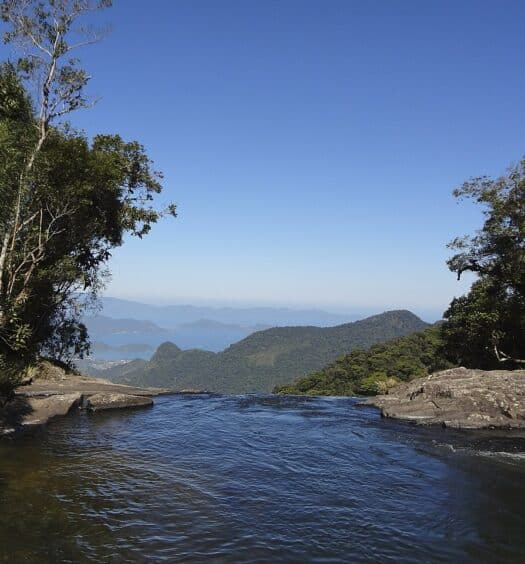 vista da Serra da Bocaina, em Bananal, com a Baía da Ilha Grande ao fundo e, à frente, um rio de águas límpidas que desemboca no que parece ser uma cachoeira, há uma margem pedregosa de ambos os lados com árvores verdes e o céu é azul