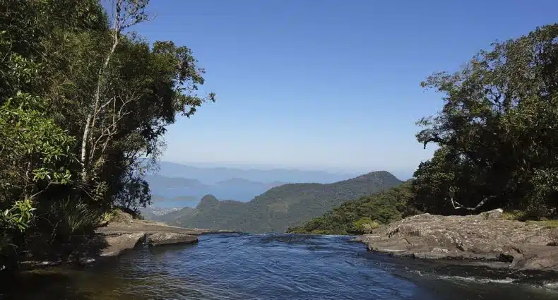 vista da Serra da Bocaina, em Bananal, com a Baía da Ilha Grande ao fundo e, à frente, um rio de águas límpidas que desemboca no que parece ser uma cachoeira, há uma margem pedregosa de ambos os lados com árvores verdes e o céu é azul