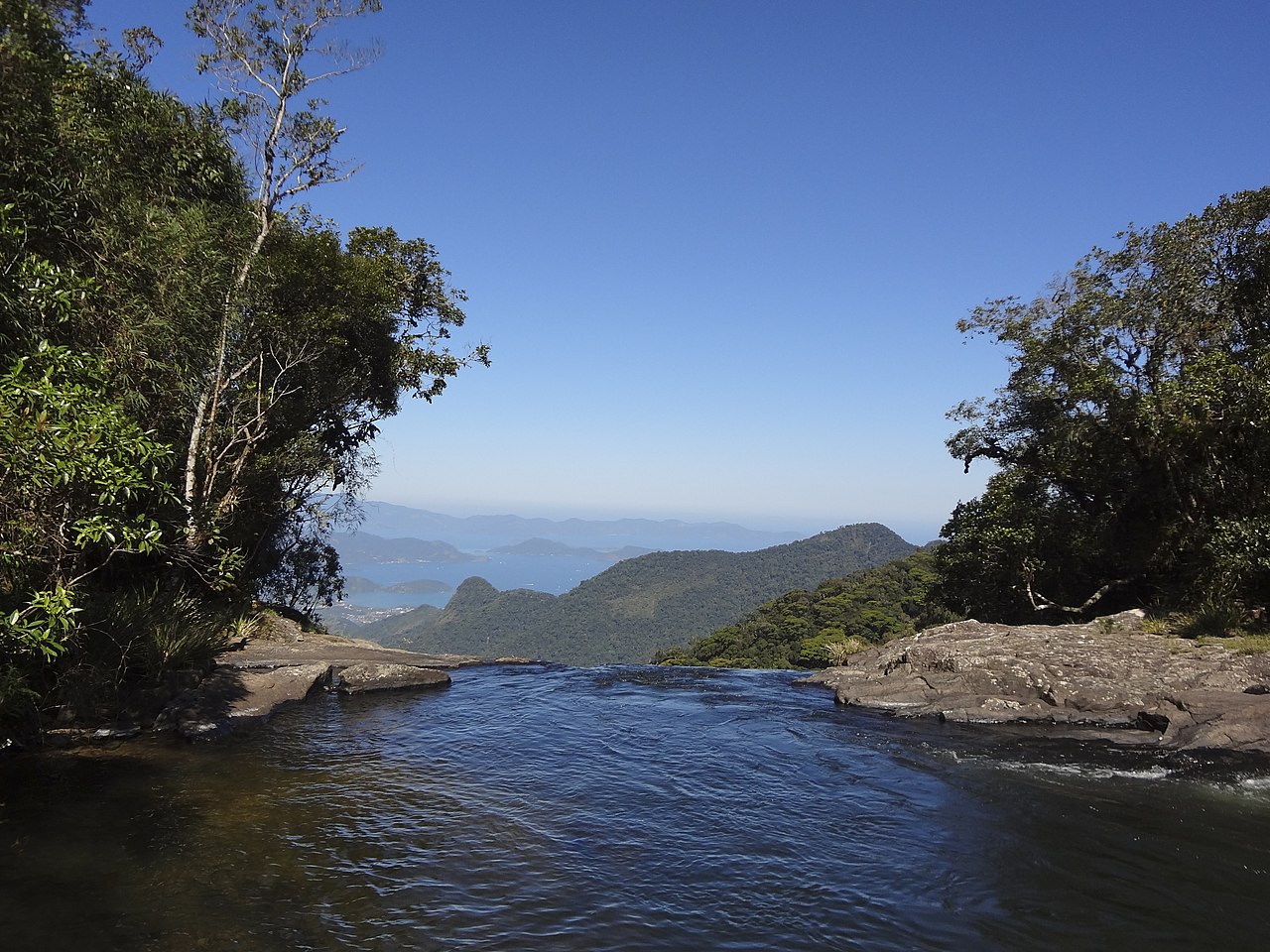 vista da Serra da Bocaina, em Bananal, com a Baía da Ilha Grande ao fundo e, à frente, um rio de águas límpidas que desemboca no que parece ser uma cachoeira, há uma margem pedregosa de ambos os lados com árvores verdes e o céu é azul