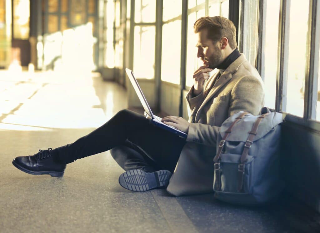 homem sentado no chão segurando um notebook no aeroporto parecendo perplexo para ilustrar o post de seguro viagem cobre cancelamento da viagem