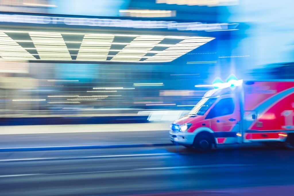 Ambulância com luzes acessas, aparentemente em alta velocidade, em frente a um hospital, numa foto de efeito de luzes que passa a impressão de estar em movimento