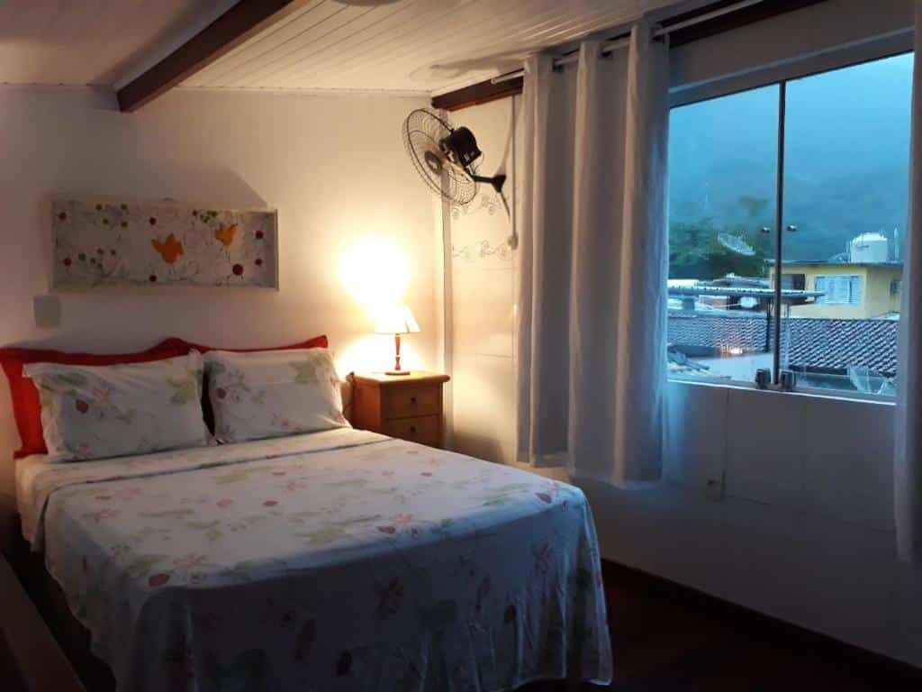 Quarto do airbnb Angel Ilha Flats e Varanda da Praia. No canto esquerdo está a cama de casal, no lado direito da cama há uma pequena cômoda com um abajur em cima e na parede do lado direito há uma janela de vidro. Imagem para ilustrar o post airbnb em Ilha Grande.