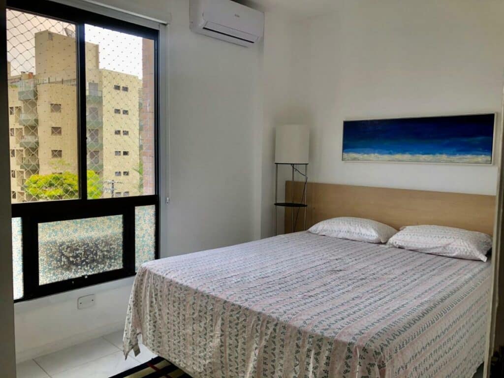 Quarto do Ap Riviera uma quadra da praia, um dos airbnb em Bertioga. Uma cama de casal com travesseiros e jogo de cama tem abajures dos dois lados e uma janela com tela na parede ao lado esquerdo. O aparelho de ar-condicionado fica ao lado da janela, para representar airbnb em Bertioga.