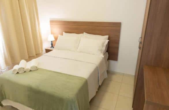 Quarto do airbnb Apart Sol Nascente. No lado direito da imagem há um armário, no lado esquerdo há uma cama de casal, no lado esquerdo da cama há uma mesa de cabeceira com um abajur em cima e na parede do lado esquerdo há uma cortina fechada. Imagem para ilustrar o post airbnb em Itatiaia.