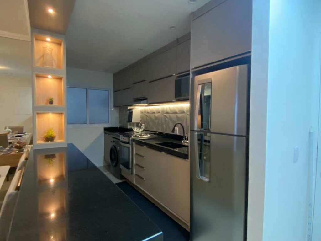 Cozinha do Apartamento em Bertioga com varanda gourmet com bancadas e armários entre a geladeira, o fogão e a máquina de lavar. Há ainda pia e micro-ondas ali.