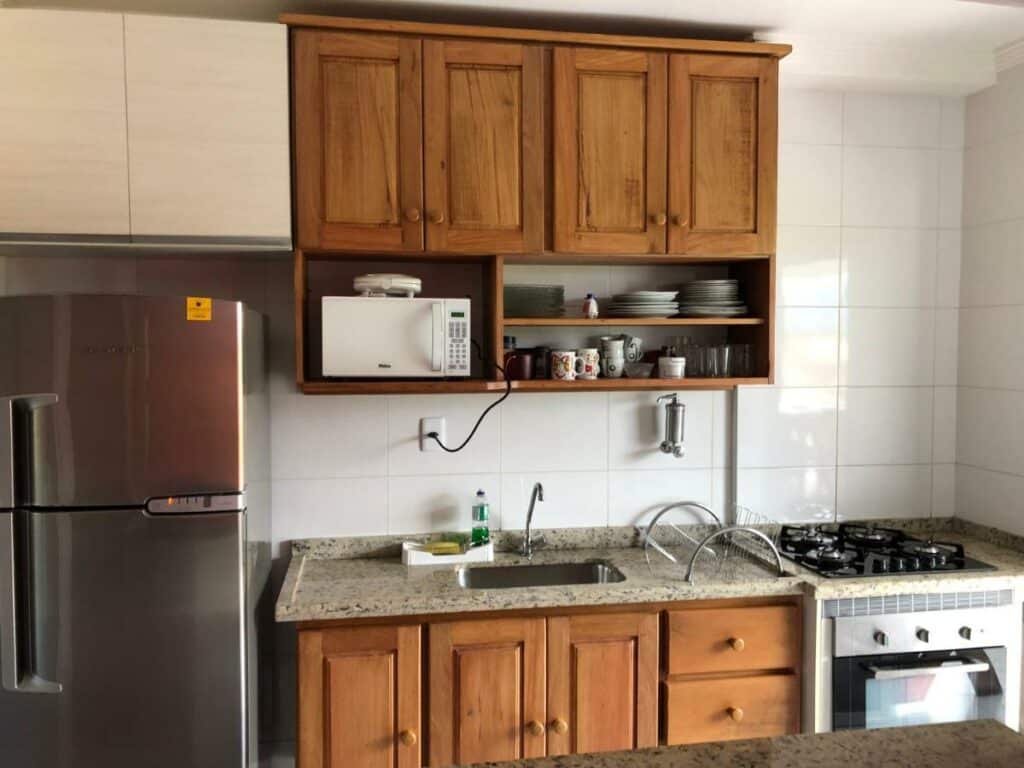 Cozinha do Apto no Indaiá Home Club, onde uma geladeira e um fogão tem bancadas e armários entre si. A pia fica logo abaixo do apoio com micro-ondas e utensílios, para representar airbnb em Bertioga.