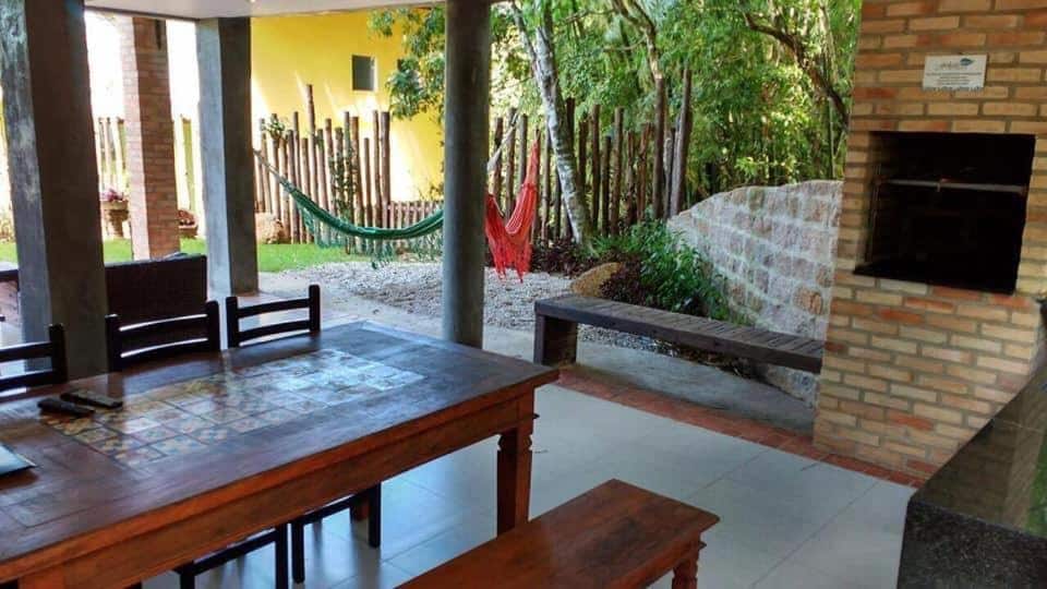 Area de lazer do Flat Felicitá Lagoinha com mesa de madeira do lado esquerdo da imagem com três cadeiras e do lado direita a churrasqueira feita de tijolo.
