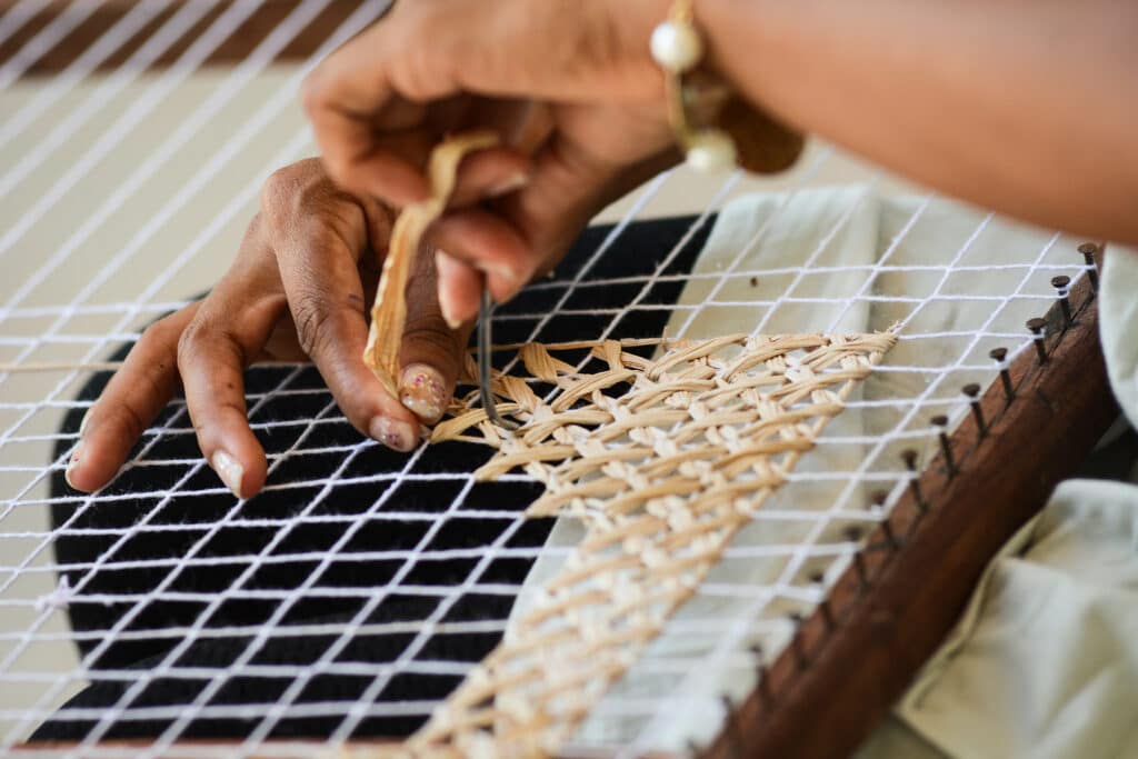 Mãos de uma mulher morena tecendo fibra feita de folha de bananeira seca. Ela está cruzando as fibras em uma tela apoiada em seu colo.