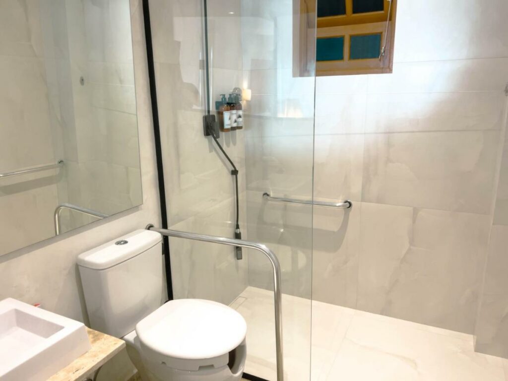 Banheiro com acessbilidade do Catalina Hotel com vaso sanitário do lado direito da imagem com barra de apoio, um box de vidro e a ao fundo o chuveiro com barra de apoio.