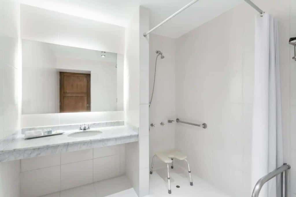 banheiro adaptado do Costa del Sol Wyndham com uma pia de mármore branca à esquerda e uma área de chuveiro à direita, bem aberta e com uma cadeira de banho