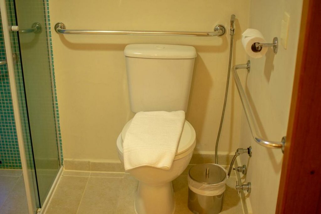 Banheiro com acessibilidade da Juquehy La Plage Hotel com vaso sanitário a frente e do lado esquerdo uma barra de apoio e ao fundo outra barra de apoio.