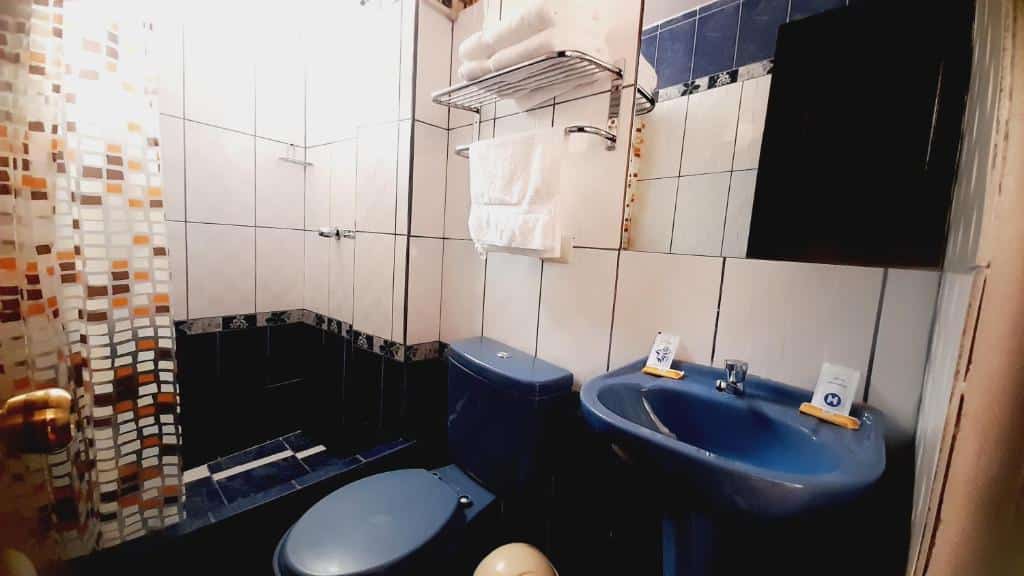 banheiro do El Tambo com louças do vaso sanitário e pia na cor azul escura, assim como o piso do chão e alguns da parede também. A ducha é separada por uma cortina em tons de laranja.