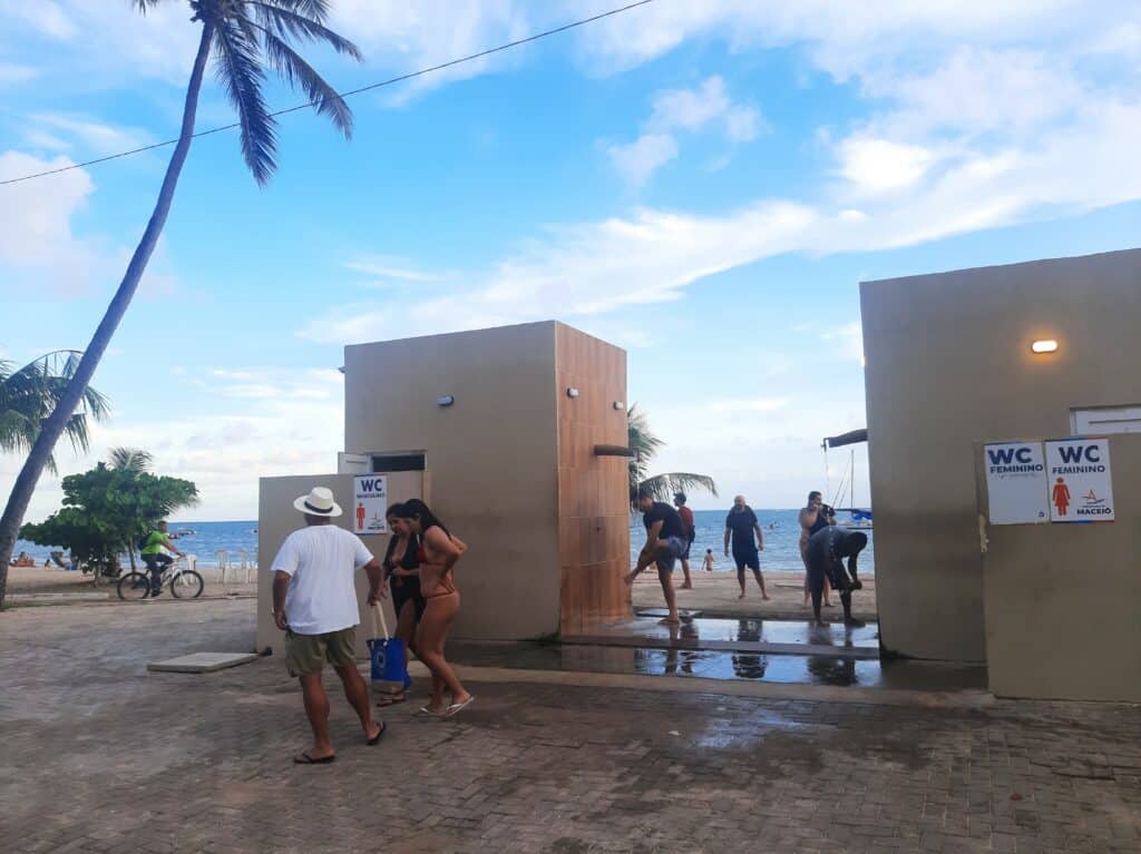 Banheiro público com pessoas se banhando na ducha e a praia de Maceió atrás, durante o dia