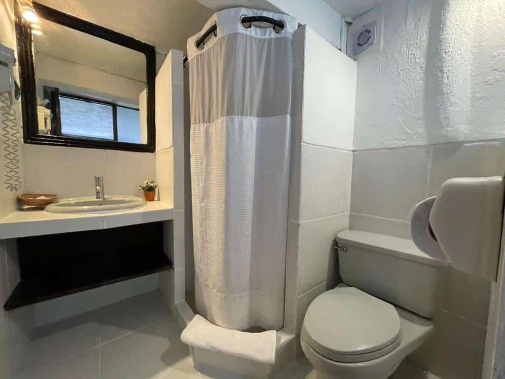 banheiro do Sacred Stone Boutique Hotel barato com um vaso sanitário à direita da imagem, uma ducha com cortina de tecido ao centro e uma pia simples de pedra com um espelho quadrado no lado esquerdo
