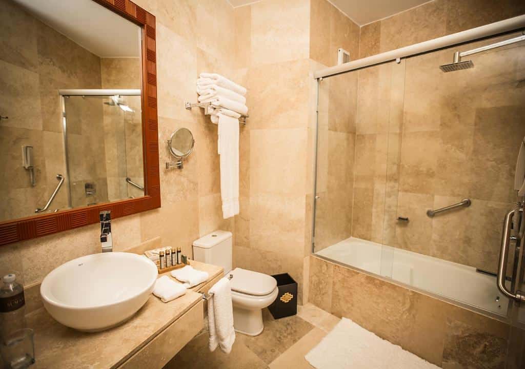 banheiro do hotel sumaq mostrando uma banheira de imersão dentro do box de vidro com ducha no lado direito da imagem, além de uma pia de pedra do lado esquerdo, com um grande espelho acima e ao lado do vaso sanitário.