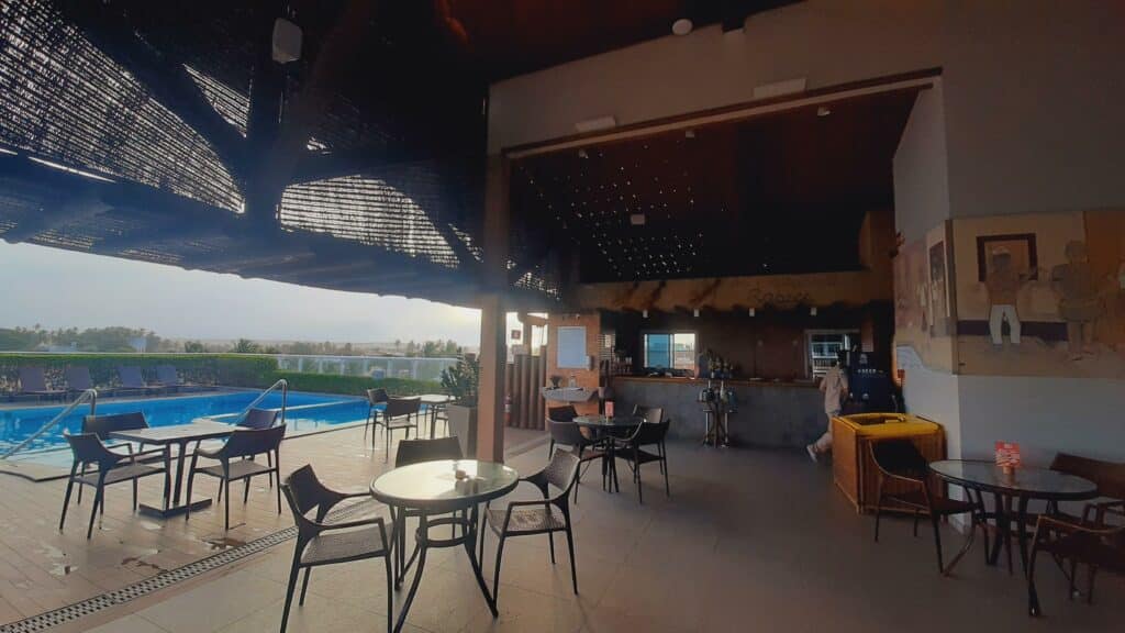 Bar localizado ao lado da piscina do hotel. Há mesas com cadeiras espalhadas, um freezer de sorvete e um balcão de madeira do bar. À esquerda fica a piscina