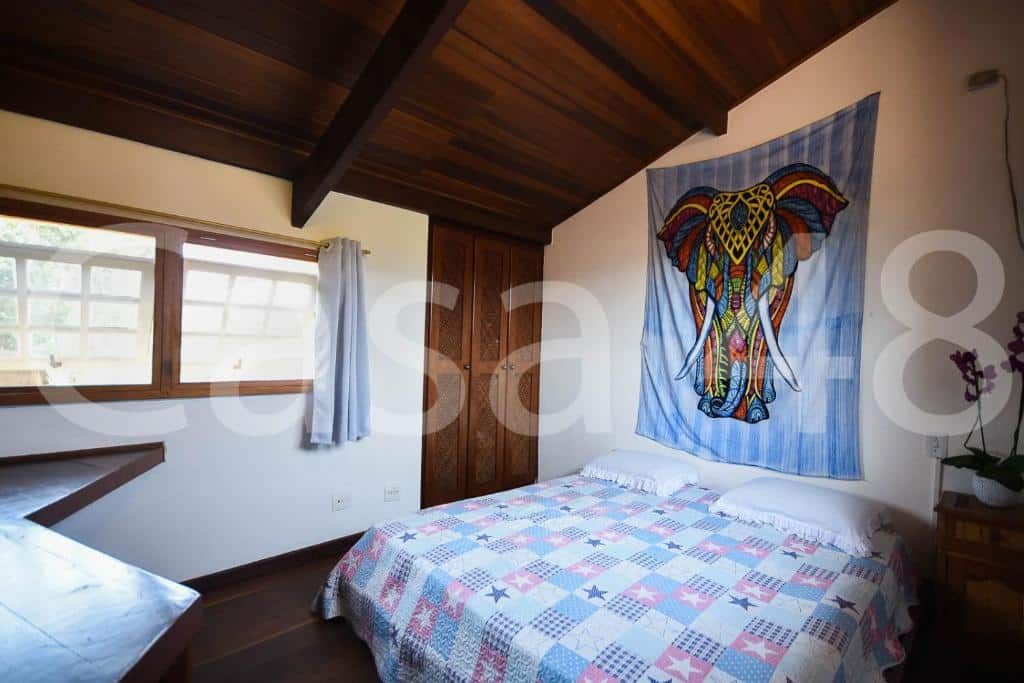 Quarto da Beach House - Camburi, um dos airbnb em Camburi. Uma cama de casal com colcha e travesseiros está encostada na parede ao lado direito e tem uma mesinha com vaso de flores ao lado, assim como um pano estampado na parede acima. Logo em frente fica uma mesa ao lado de uma janela.