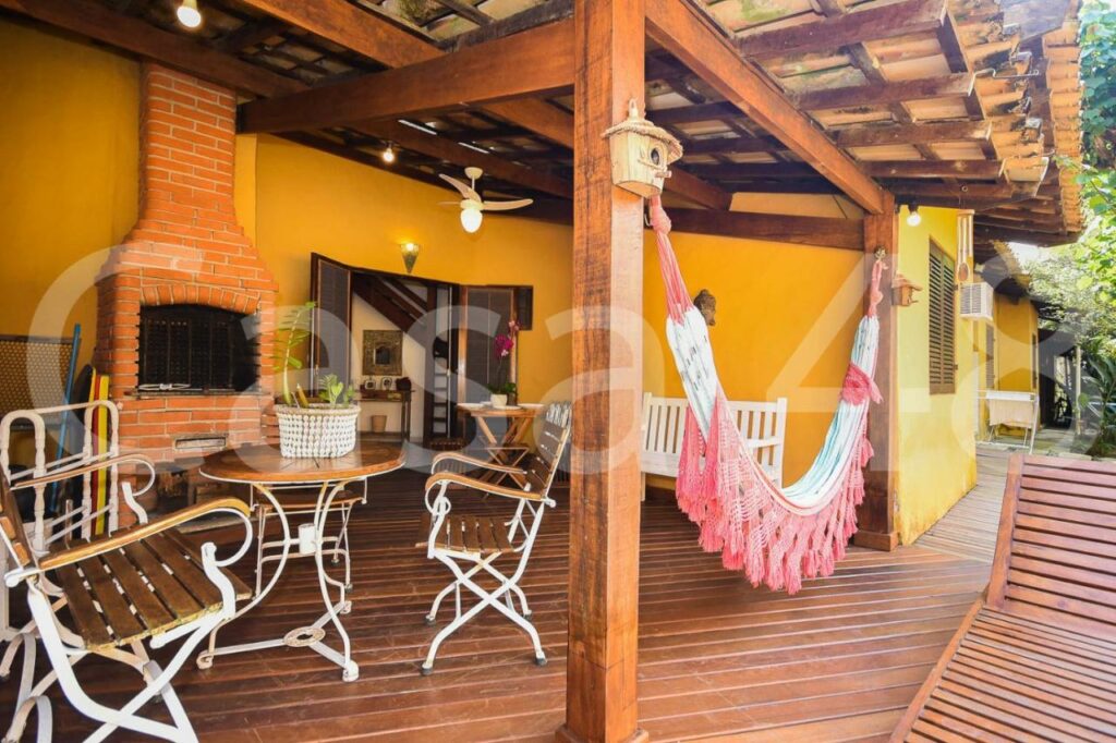 Varanda de madeira da Beach House - Camburi, onde há uma rede, uma churrasqueira, uma espreguiçadeira e uma mesinha com cadeiras.