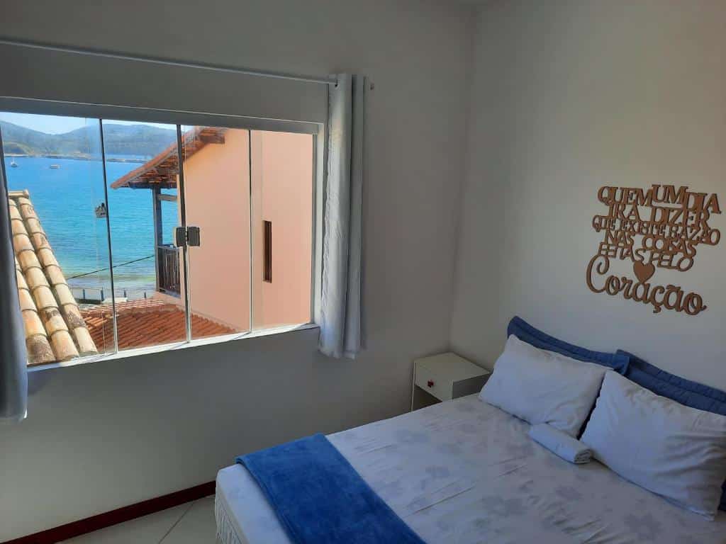 Quarto do airbnb Bela Vista Flats. A cama de casal está encostada na parede do lado direito da imagem e no lado esquerdo da cama há uma pequena mesa de cabeceira. Ainda no lado esquerdo há uma janela de vidro com cortinas e vista para o mar. Imagem utilizada para ilustrar o post airbnb em Arraial do Cabo.