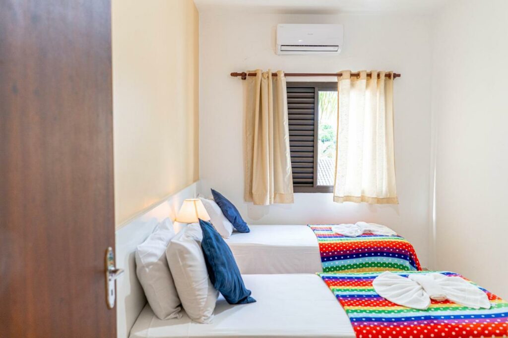 Quarto da Bella Casa Flat Service, um dos airbnb em Bertioga. Duas camas de solteiro com travesseiros e jogo de cama estão encostadas na parede ao lado esquerdo. Ao fundo há uma janela com cortinas e um aparelho de ar-condicionado logo acima.