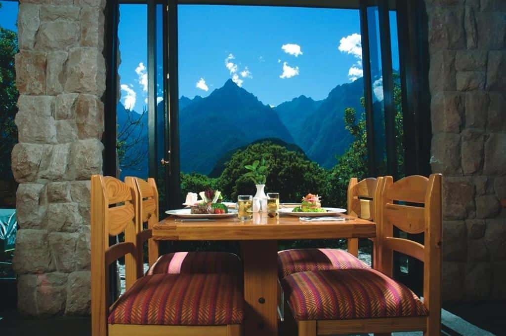 restaurante do Belmond Sanctuary Lodge com uma mesa de madeira retangular com quatro cadeiras, alguns pratos com alimentos em cima e uma ampla janela com vista para as montanhas logo atrás da mesa.