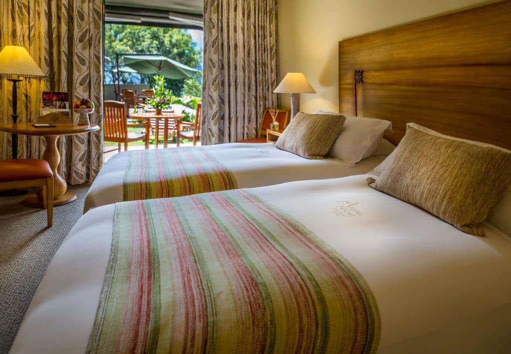 quarto duplo do Belmond Sanctuary Lodge para representar onde ficar em Machu picchu, com duas camas de solteiro no lado direito da imagem, e um porta dupla aberta ao fundo do quarto, dando acesso a varanda e ao jardim do hotel.