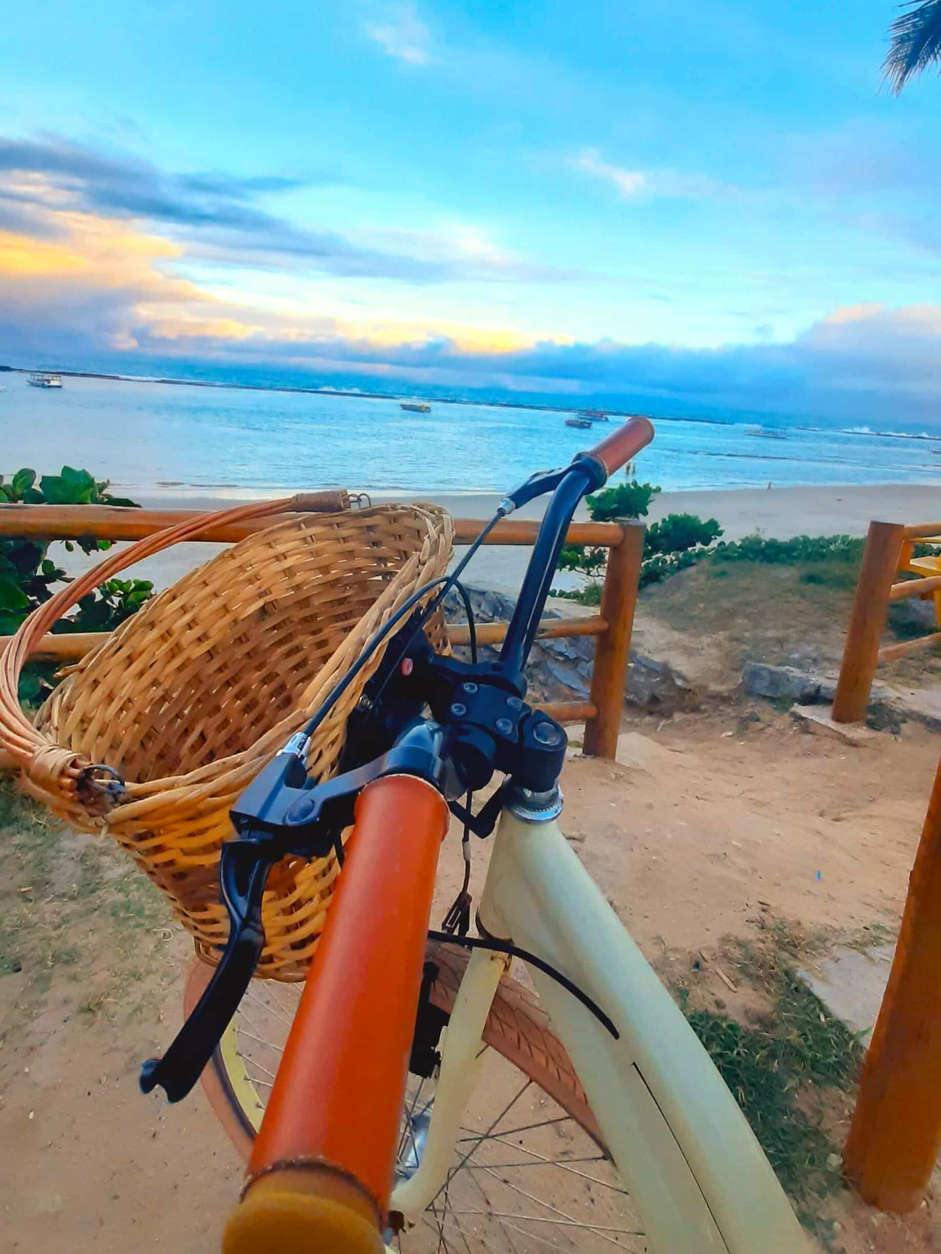 Guidão de uma bicicleta com cestinha inclinado com o pôr de sol na praia do Francês ao fundo