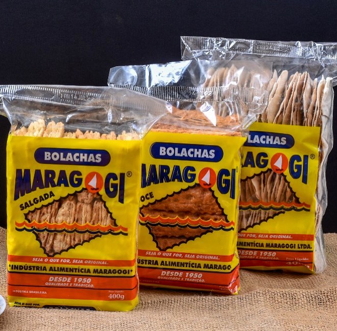 Três pacotes de bolacha em cima de uma mesa com o fundo preto. As bolachas tem pacote amarelo escrito "Bolachas Maragogi; Seja o que for, seja original; indústria alimentícia Maragogi; desde 1950 qualidade e tradição; industria brasileira 400g. Há pacotes de bolacha doce e salgada