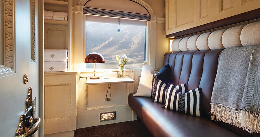 vagão dormitório do andean explorer com um sofá-cama à direita, uma janela com vista para as montanhas, uma mesa com abajur e um arranjo de flores abaixo da janela e uma porta semiaberta à esquerda com maçaneta dourada e elegante