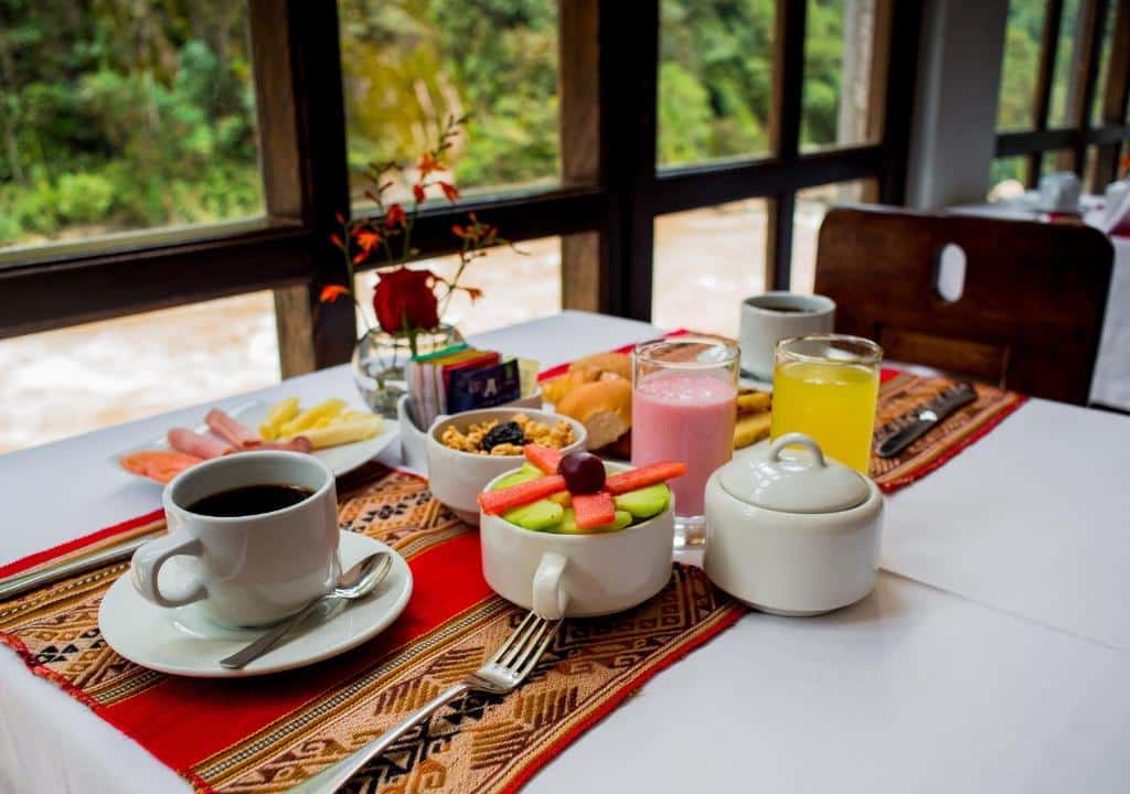 mesa de café da manhã do hatun inti boutique hotel bem ao lado da janela que dá vista ao rio. Na mesa há uma xícara de café, copos de suco e iogurte, frutas e enroladinhos de queijo e presunto.
