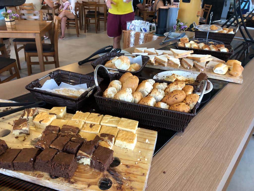 Café da tarde servido no Maragogi Brisa Exclusive Hotel, sendo uma mesa com travessas contendo bolos cortados, paães e torradas