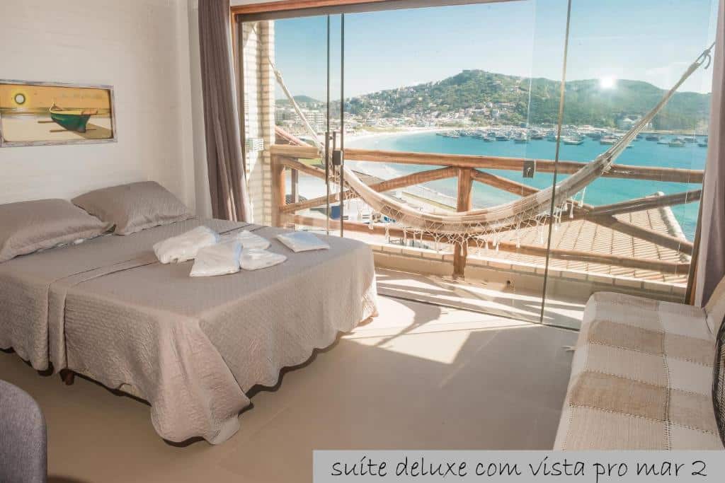 Quarto do airbnb Cantinho dos Anjos Suítes. A cama de casal está encostada na parede do lado esquerdo da imagem, na frente da cama há um sofá e ao fundo há uma grande porta de vidro que dá acesso a varanda. É possível ver uma rede de descanso na varanda e também há vista para a praia. Imagem utilizada para ilustrar o post airbnb em Arraial do Cabo.