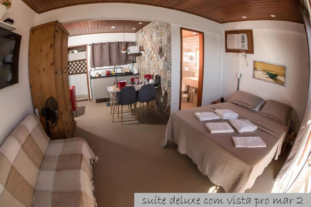 Imagem do airbnb Cantinho dos Anjos Suítes. No lado direito da imagem há uma cama de casal encostada na parede, em frente há cama há um sofá e em cima do sofá há uma TV presa a parede e no lado esquerdo da cama há uma porta que dá acesso ao banheiro. No fundo do cômodo há uma mesa de jantar com cadeiras, um frigobar e uma pia. Ainda no fundo há um armário e uma porta.