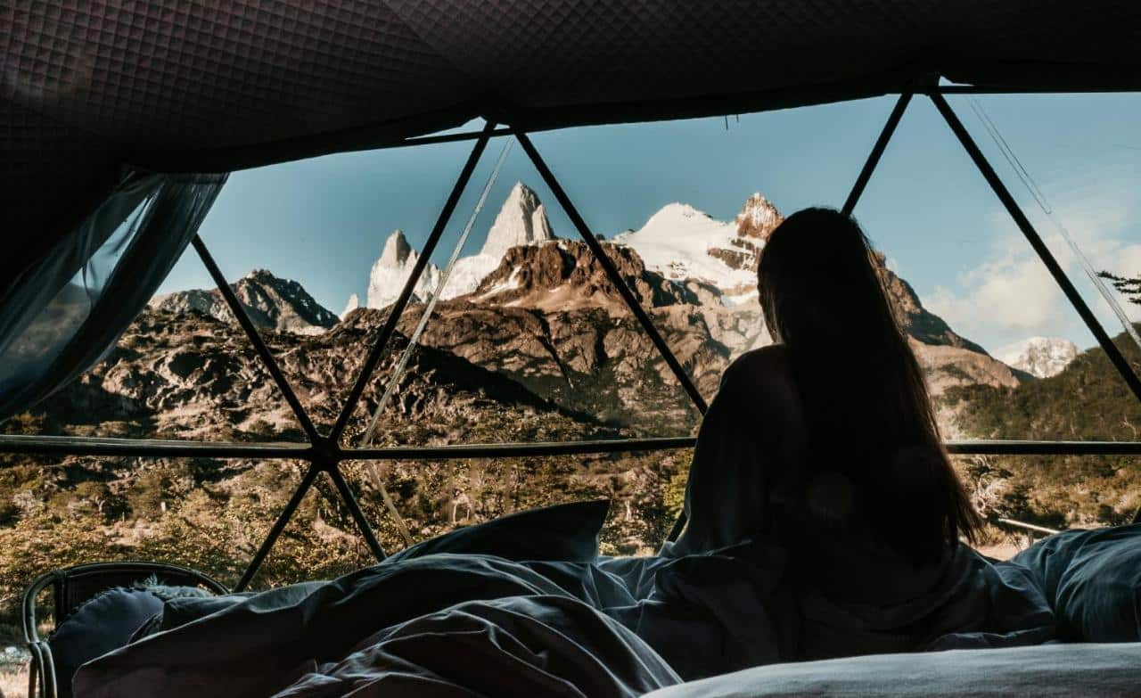 Mulher dentro do quarto do Patagonia Eco Domes, durante o dia vendo as montanhas em uma janela panorâmica. Representa hotéis em El Chalten.