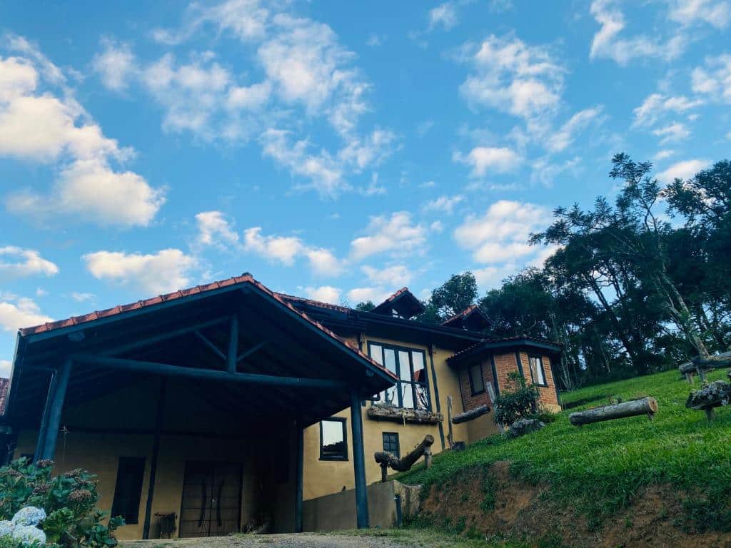 Imagem do exterior do Chalé SPAço Alto das Serra, em SFX. O céu está azul e com nuvens, o chalé está no centro da foto, em meio ao gramado. Possui várias janelas e um telhado cobrindo a entrada.