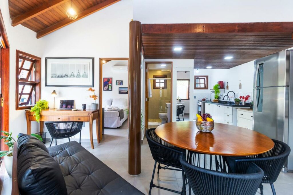 Visão geral da Casa Charmosa com Piscina em Barra do Una. A foto foi tirada ao lado de um sofá de couro que encara uma mesa redonda com três cadeiras. Ao lado esquerdo do sofá fica uma mesa de trabalho com computador, abajur, vaso de flores e cadeira. Ao lado é possível ver um pedaço da cama na porta aberta que leva ao quarto. Ao fundo da foto, depois da mesa redonda, fica a cozinha com geladeira, armários, fogão e mais utensílios. Ao lado está o banheiro.