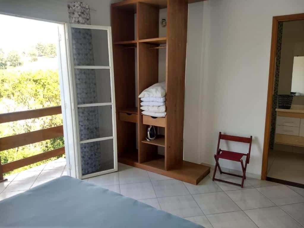 Foto do quarto em Casa com linda Vista no Centro. A cama está no canto inferior esquerdo vemos parte do pé do colchão. De frente há uma estante de madeira com almofadas, e de seu lado esquerdo há uma varanda. Na direita há uma porta para o banheiro.
Ilustra o post sobre airbnb em Serra Negra.