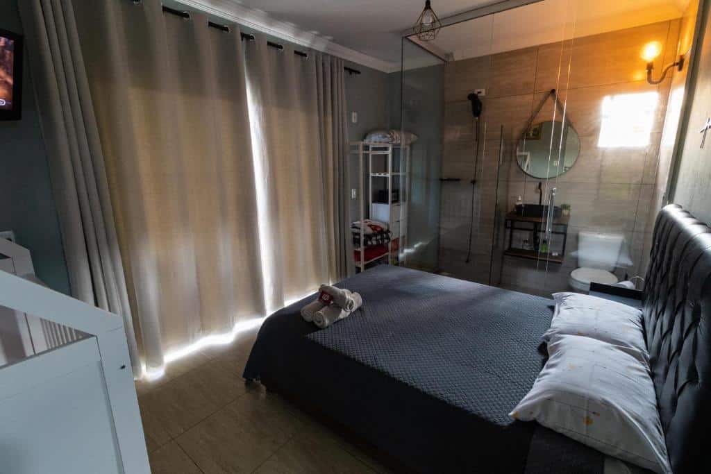 Foto do quarto da Casa com Linda Vista e Piscina. Ilustra o post sobre airbnb em Ouro Preto. Vemos a imagem de cima. A cama é box e de casal e está na direita. Na frente há uma janela grande com cortinas. Do lado da cama há um box de vidro com uma pia, vaso e chuveiro dentro. 