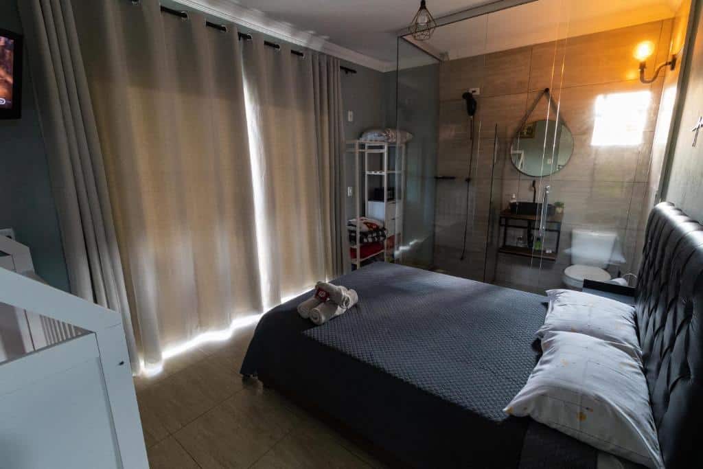 Foto do quarto da Casa com Linda Vista e Piscina. Ilustra o post sobre airbnb em Ouro Preto. Vemos a imagem de cima. A cama é box e de casal e está na direita. Na frente há uma janela grande com cortinas. Do lado da cama há um box de vidro com uma pia, vaso e chuveiro dentro.