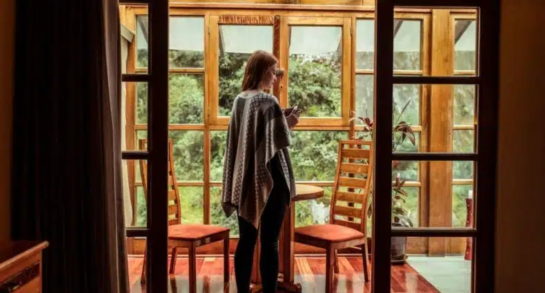 varanda de uma suíte do hotel Casa del Sol em Machupicchu com uma mulher ruiva, de cabelo longo, parada em pé no centro da imagem, usando um poncho típico da região, segurando uma xícara com as duas mãos. Ao fundo é possível ver bastante vegetação verde, do lado de fora da varanda de madeira.