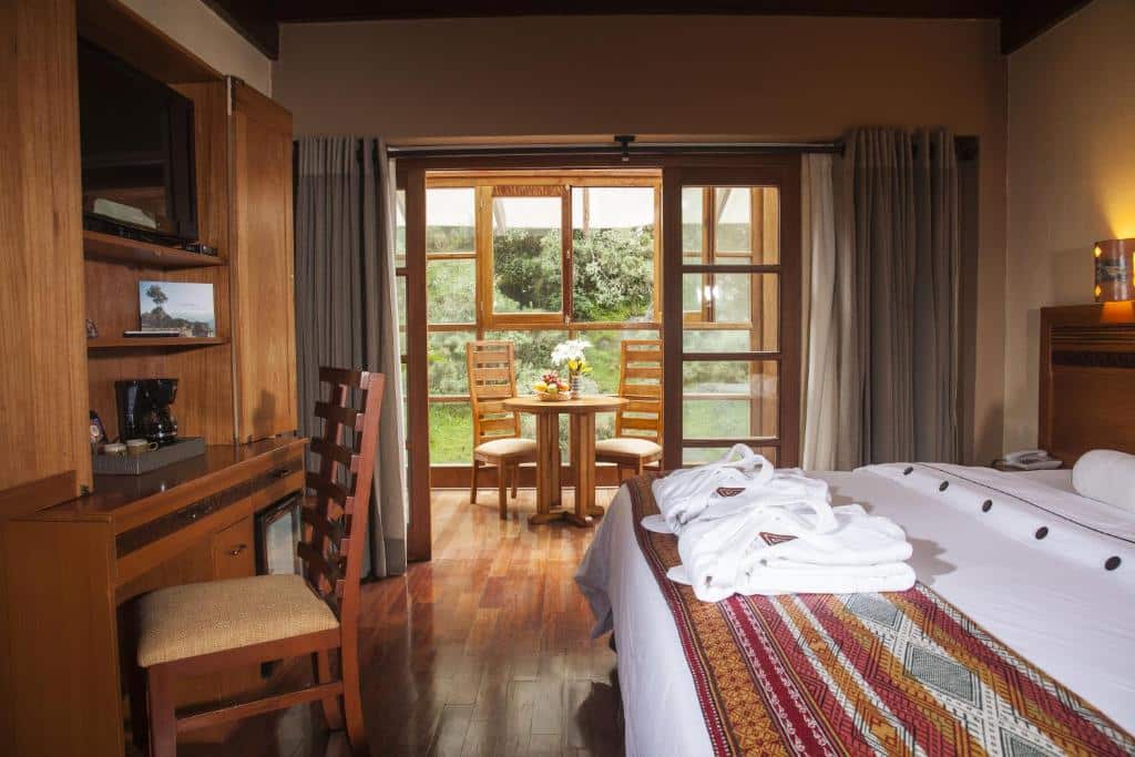 suíte da casa del sol machupicchu com uma cama de casal à direita da imagem, com dois roupões brancos dobrados aos pés da cama, uma mesa de madeira em frente a cama com uma cadeira e uma televisão pendurada acima, na parede. ao fundo é possível ver uma varanda particular com uma mesa de madeira e duas cadeiras, além de uma vista para a vegetação